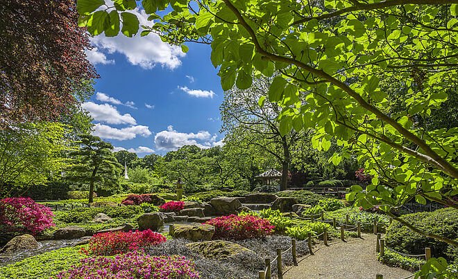 Bunte Blumen, ein Teich mit einer Statue und Bäumen. Im Hintergrund ist ein Pavillon.