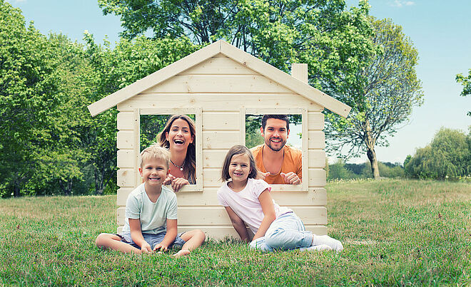 Zwei Kinder sitzen auf einer Wiese, im Hintergrund schauen eine Frau und ein Mann aus einem Spielzeughaus, Weiterverlinkung zur swa Hausanschluss-Seite