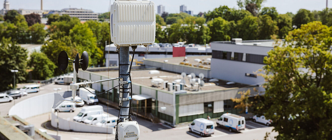 LoRaWAN-Mast im Hintergrund sind Gebäude und Bäume. 