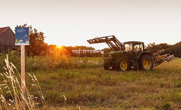 Ein Traktor auf einer Wiese. Links ist eine Scheune. Im Hintergrund sieht man den Sonnenuntergang.