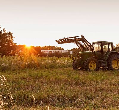 Ein Traktor auf einer Wiese. Links ist eine Scheune. Im Hintergrund sieht man den Sonnenuntergang.