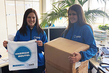 Zwei Frauen mit blauen swa Jacken stehen in einem Büro. Die Frau links hält eine Tasche der swa mit dem Aufdruck 