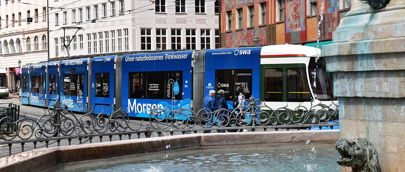 Bedruckte Straßenbahn mit swa Branding. Im Vordergrund sind Leute und ein Brunnen