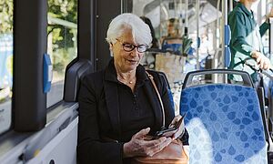 Ältere Dame mit Brille die im Bus sitzt und auf ihr Smartphone schaut. Mit Link zum öffnen einen Pop-Ups mit Informationen zum Mobil-Abo 9 Uhr