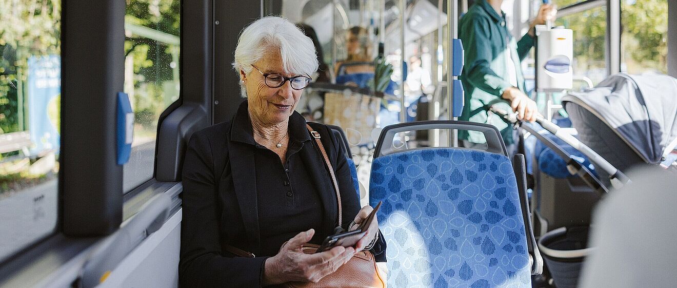 Ältere Dame mit Brille die im Bus sitzt und auf ihr Smartphone schaut. Mit Link zum öffnen einen Pop-Ups mit Informationen zum Mobil-Abo 9 Uhr