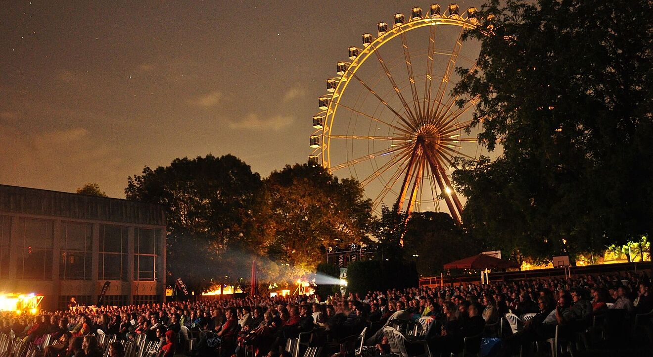 Viele Personen auf Stühlen bei der swa Kurzfilmnacht 2024. Im Hintergrund sind Bäume und ein Riesenrad
