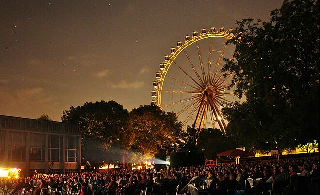 Viele Personen auf Stühlen bei der swa Kurzfilmnacht 2024. Im Hintergrund sind Bäume und ein Riesenrad