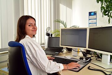 Eine junge Frau sitzt am Schreibtisch vor einem Computerbildschirm. Sie trägt ein Headset. Zudem sind Blätter einer Büropflanze zu sehen.