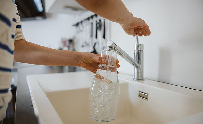 Person hält eine Karaffe unter einen Wasserhahn und befüllt diese mit Leitungswasser