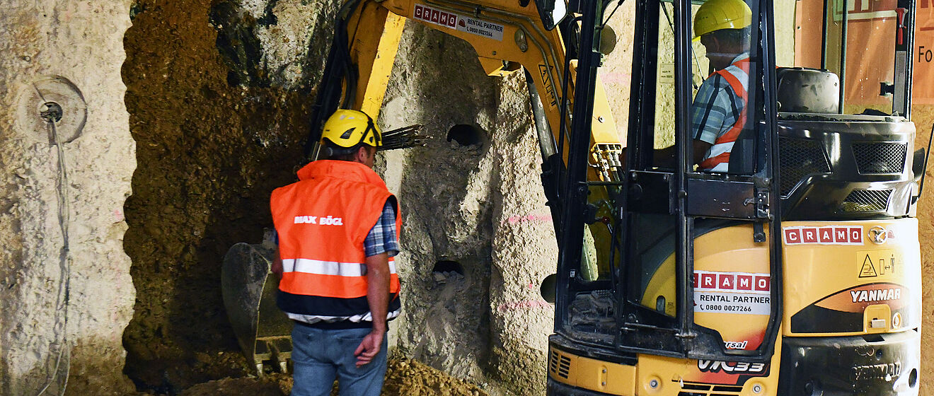Zwei Bauarbeiter in Schutzkleidung, die in einer felsigen, unterirdischen Umgebung einen gelben Minibagger bedienen.