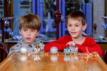 Zwei Jungen sitzen in der weihnachtlich geschmückten Christkindltram der Stadtwerke Augsburg (swa). Der Holztisch ist mit glänzenden silbernen und blauen Christbaumkugeln und einer Lichterkette dekoriert.
