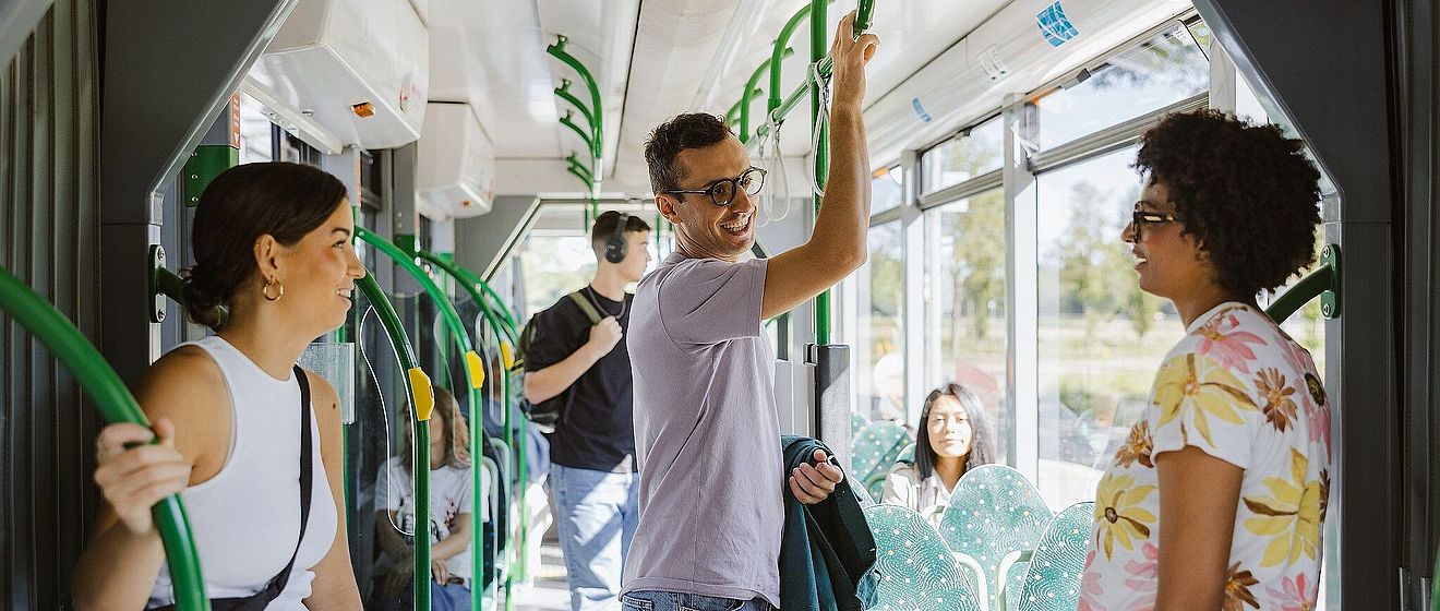 Mann mit Brille lacht und steht in der Straßenbahn. Links und rechts von ihm sind zwei Frauen, die sich an einer Stange festhalten. Im Hintergrund sind weitere Fahrgäste.
