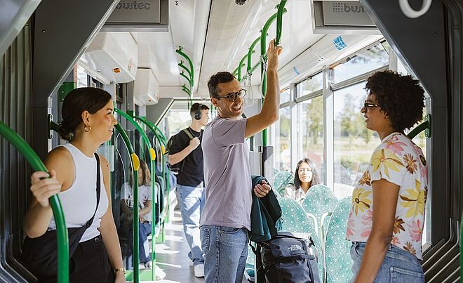 Mann mit Brille lacht und steht in der Straßenbahn. Links und rechts von ihm sind zwei Frauen, die sich an einer Stange festhalten. Im Hintergrund sind weitere Fahrgäste.