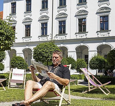 Mann sitzt auf Liegestuhl und ließt eine Zeitung, im Hintergrund sind weitere Liegestühle und ein Gebäude zu sehen.