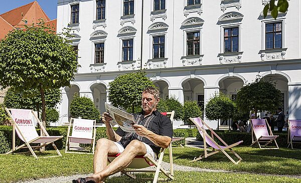 Mann sitzt auf Liegestuhl und ließt eine Zeitung, im Hintergrund sind weitere Liegestühle und ein Gebäude zu sehen.