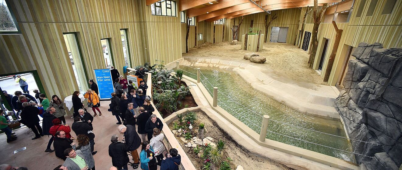 Eine große Gruppe von Menschen nimmt an einem feierlichen Eröffnungs-Event im neuen Elefantenhaus des Augsburger Zoos teil, das durch seine moderne Architektur mit Holzbalken und naturgetreuer Umgebung besticht.