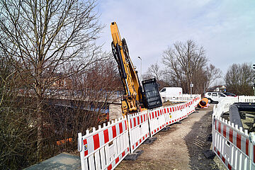 Bagger fährt auf einer Baustelle an der Wertachbrücke. Links und im Hintergrund sind Bäume. Ein Klick sorgt für eine Großansicht des Bildes.