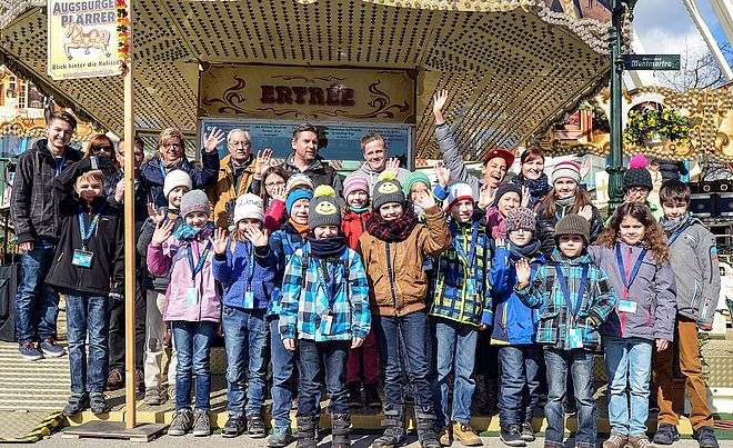 Gruppe von Kindern und Erwachsenen lächelt und winkt in die Kamera.