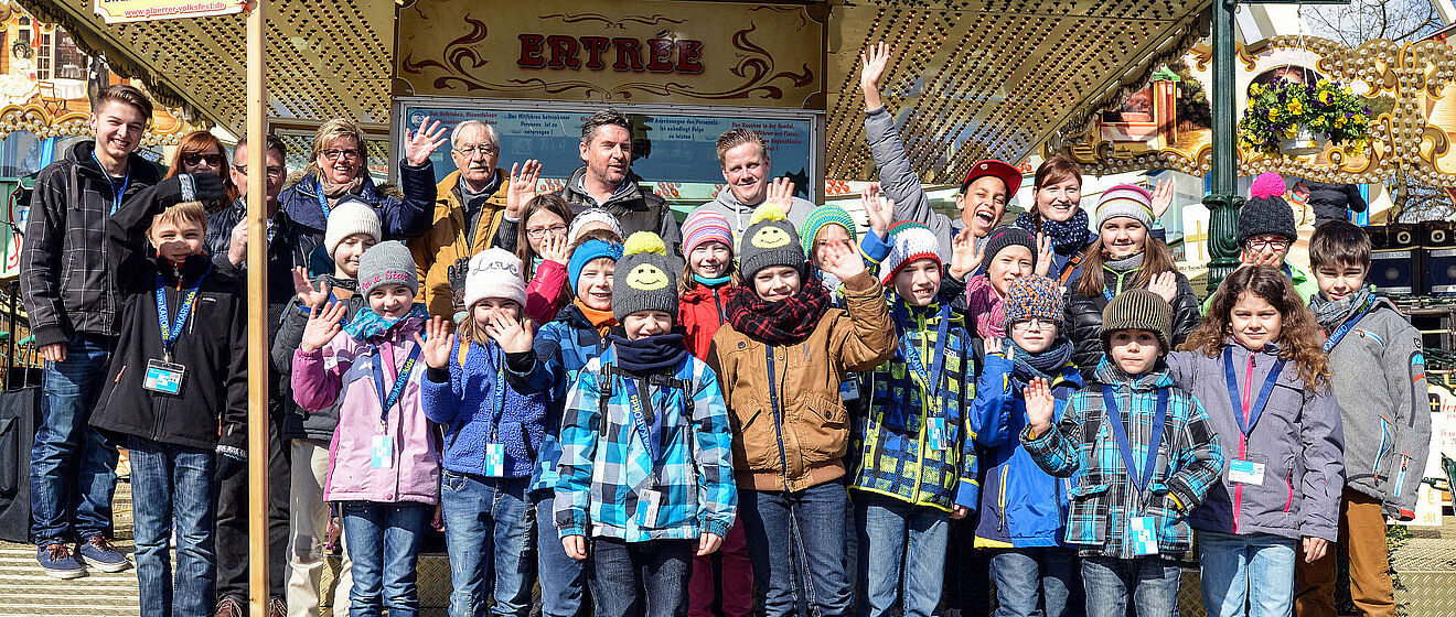 Gruppe von Kindern und Erwachsenen lächelt und winkt in die Kamera.