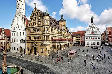 Marktplatz mit Menschen einem Brunnen und Gebäuden im Hintergrund in der Stadt Rothenburg ob der Tauber. Ein Klick sorgt für eine Großansicht des Bildes.