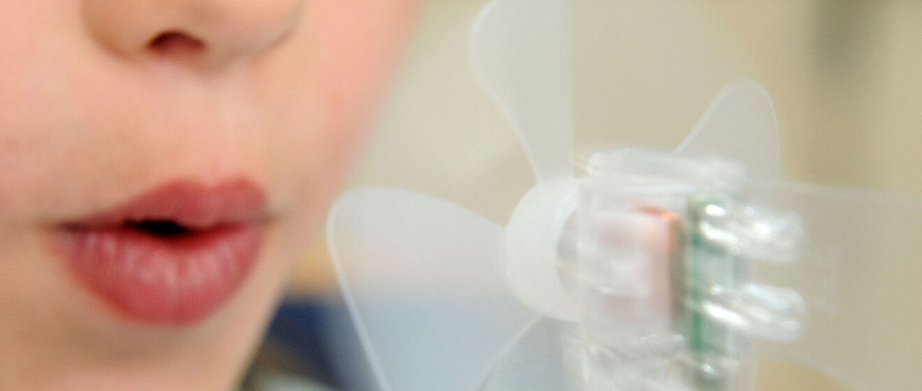 Energie wird erklärt. Ein Mund pustet gegen ein Modellwindrad.