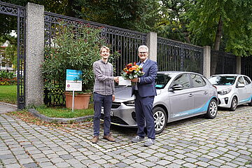 Zwei Männer schütteln die Hände vor einem Carsharing Auto. Der rechte Mann hält einen Blumenstrauß