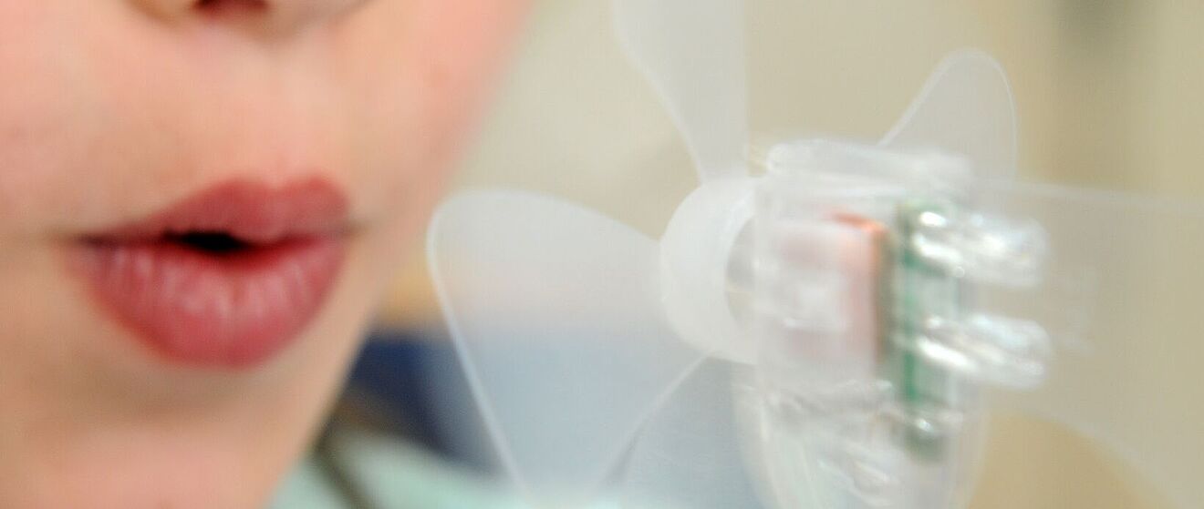 Eine Person bläst in einen kleinen, transparenten Mini-Ventilator, dessen Rotorblätter sich durch den Luftstrom drehen.