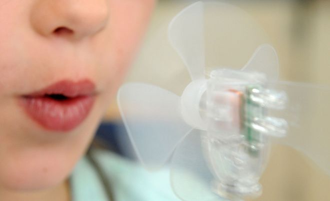 Eine Person bläst in einen kleinen, transparenten Mini-Ventilator, dessen Rotorblätter sich durch den Luftstrom drehen.
