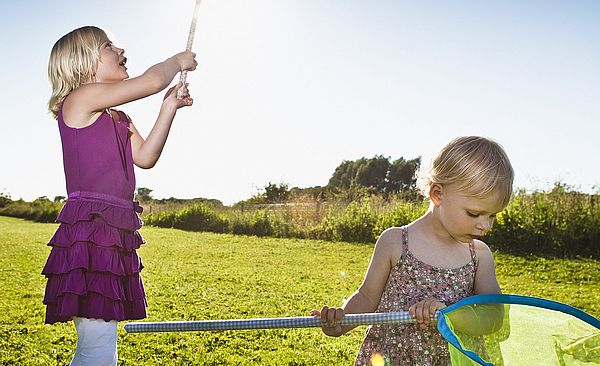 Zwei Kinder spielen mit Keschern auf einer Wiese. Die Sonne scheint.