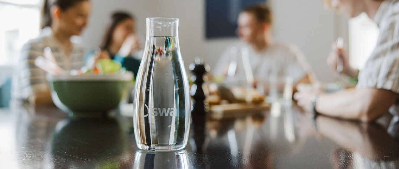 Im Fokus ist eine mit Wasser gefüllte swa Karaffe. Im Hintergrund sind Personen an einem Esstisch am Essen