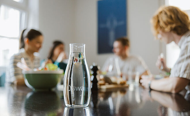 Im Fokus ist eine mit Wasser gefüllte swa Karaffe. Im Hintergrund sind Personen an einem Esstisch am Essen