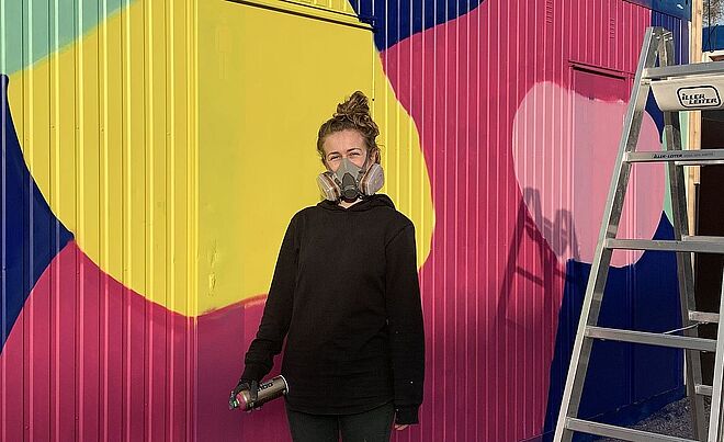 Frau mit Atemschutzmaske und Spraydose steht vor einem bemalten WC Container. Weiterverlinkung auf WC Container Newsartikel