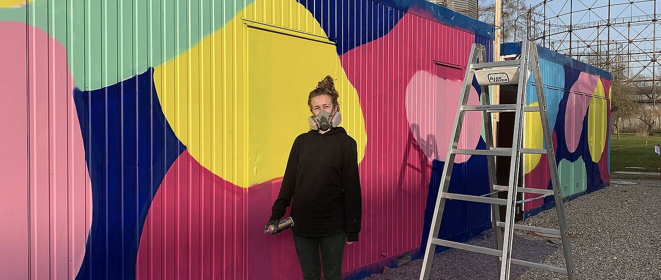 Frau mit Atemschutzmaske und Spraydose steht vor einem bemalten WC Container. Weiterverlinkung auf WC Container Newsartikel
