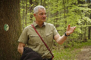 Förster steht im Wald und erklärt dem Kamerateam etwas. - Ein Klick auf das Bild führt zur vergrößerten Fotoansicht.