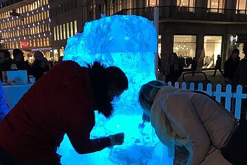 Zwei Menschen, ein Mann in einer roten Jacke und eine Frau in einem hellen Mantel, beugen sich über einen großen, intensiv blau beleuchteten Eisblock, der einen belebten Stadtplatz bei Nacht dominiert. Umgeben von städtischen Lichtern und Passanten, scheinen sie das Innere des Eises zu bearbeiten. Ein Klick auf das Bild führt zu einer größeren Ansicht.