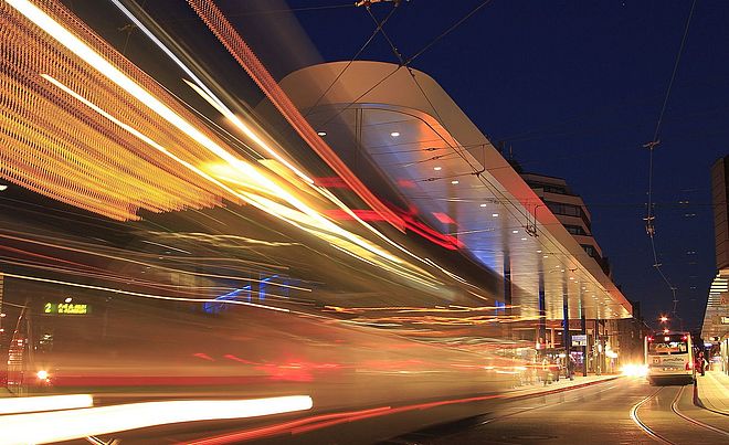 Haltestelle am Königsplatz bei Nacht. Links befindet sich die verschwommene Silhouette einer vorbeifahrenden Straßenbahn.