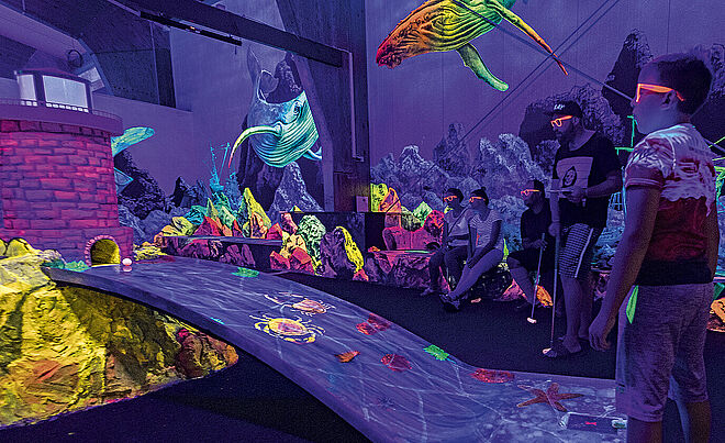 Personen mit Neonbrille beim Neon Minigolf schauen auf einen Minigolfball.