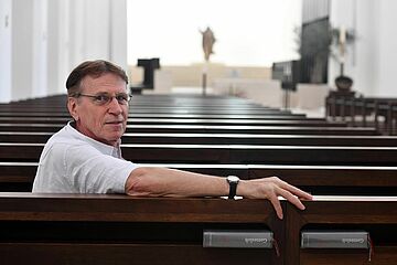 Mann sitzt auf Bank in der Kirche und lächelt in die Kamera - Ein Klick auf das Bild führt zur vergrößerten Fotoansicht.