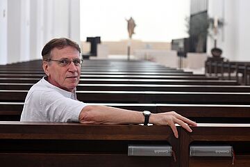 Mann sitzt auf Bank in der Kirche und lächelt in die Kamera. - Ein Klick auf das Bild führt zur vergrößerten Fotoansicht.