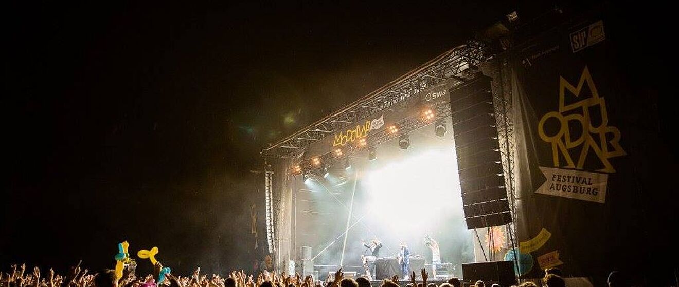 Ein LIVE-Musik-auftritt an einem vergangenen Modular-Festival in Augsburg. Die Bühne wird von Lichtern bestrahlt und eine große Menge von Menschen stehen davor.