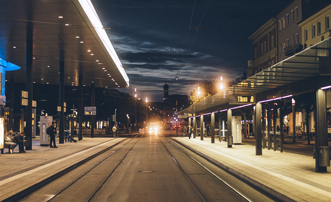 Eine modern gestaltete Straßenbahnhaltestelle bei Nacht, mit langen, beleuchteten Überdachungen über den Gleisen. Im Hintergrund ist ein beleuchtetes Hochhaus und eine Stadtansicht mit weiteren Gebäuden zu erkennen.
