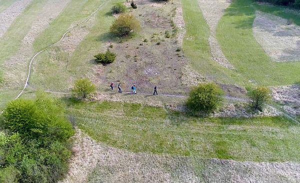 Luftansicht einer Landschaft. Inmitten dieser laufen mehrere Personen entlang eines Wanderwegs.
