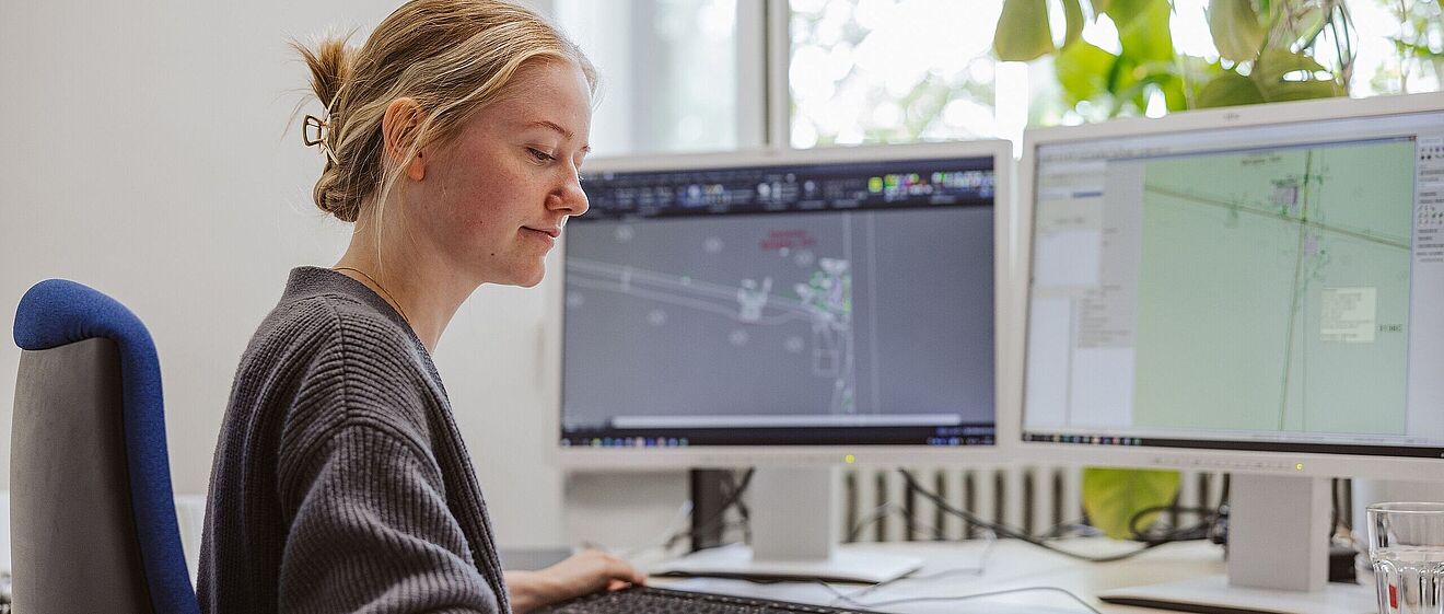 Frau sitzt auf einem Bürostuhl am Schreibtisch vor zwei Monitoren