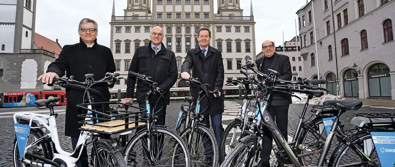 Die beiden Geschäftsführer der swa Alfred Müllner und Dr. Walter Casazza, der Oberbürgermeister Dr. Kurt Gribl sowie ein weiterer Mann stehen in dunkler Kleidung mit eBikes der swa auf dem Augsburger Rathausplatz. Im Hintergrund befindet sich der Perlachturm, das Rathaus sowie zwei Straßenbahnen.
