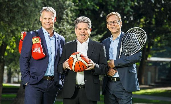 Drei Männer stehen mit Boxhandschuhen, einem Basketball und einem Tennisschläger draußen vor Bäumen.