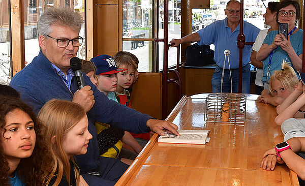 Der Geschäftsführer der swa sitzt mit einem Mikrofon umgeben von Kindern vor einem Buch in einer historischen Tram. Im Hintergrund sind weitere Erwachsene, von denen eine Frau ein Foto macht.