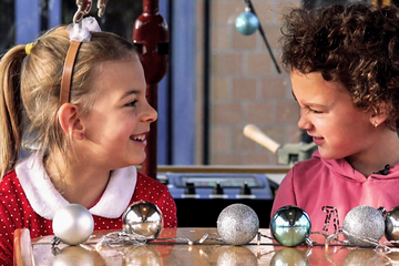 Zwei Mädchen sitzen in der weihnachtlich geschmückten Christkindltram der Stadtwerke Augsburg (swa) und gucken sich gegenseitig an. Der Holztisch ist mit glänzenden silbernen und blauen Christbaumkugeln dekoriert.