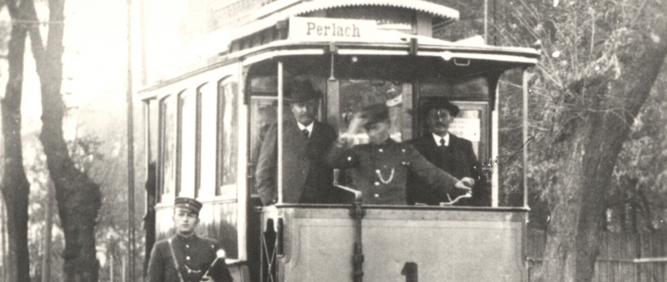 Historische Aufnahme einer alten Tram mit Fahrer und zwei Fahrgästen.