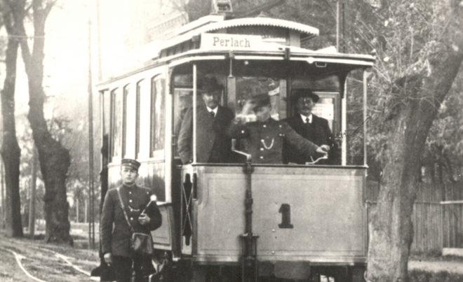 Historische Aufnahme einer alten Tram mit Fahrer und zwei Fahrgästen.