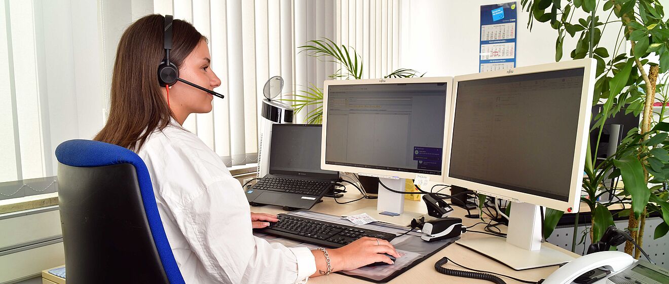 Eine junge Frau sitzt am Schreibtisch vor einem Computerbildschirm. Sie trägt ein Headset. Zudem sind Blätter einer Büropflanze zu sehen.